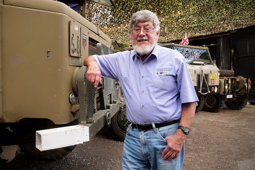 Norm Austin leans on a military vehicle with other trucks in the background.
