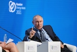Vladimir Putin sitting on a chair on a stage against a blue background.