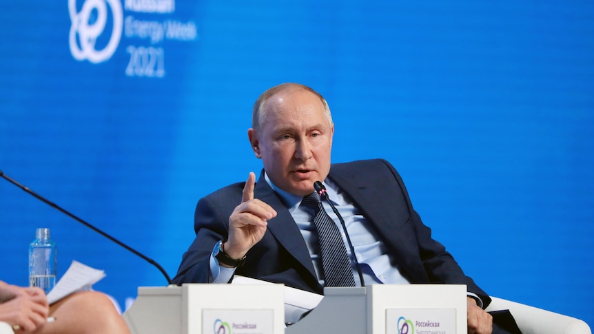 Vladimir Putin sitting on a chair on a stage against a blue background.