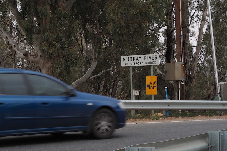 A car drives over a low bridge, flanked by gum trees and adorned with a sign reading Abbotsford Bridge