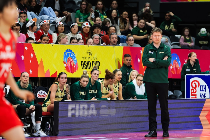 Opals coach Shannon Seebohm stands on the sideline with his arms folded, players sit on a bench behind him.