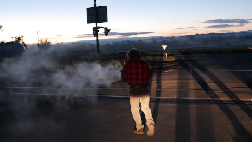  man in a flannel shirt performs a smoking ceremony on the edge of a train line in low dawn light.