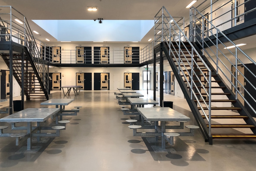 Steel tables are bolted to the floor in a large room with a second storey walkway. Identical doors to cells line the two floors.