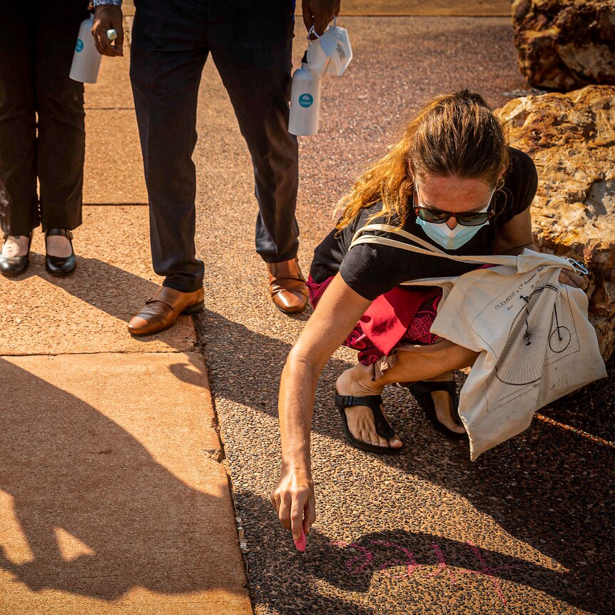 Woman in mask kneeling and chalking 'tree' on the ground while people watch.