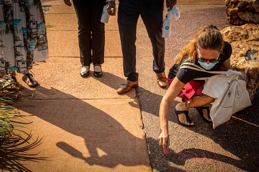 Woman in mask kneeling and chalking 'tree' on the ground while people watch.