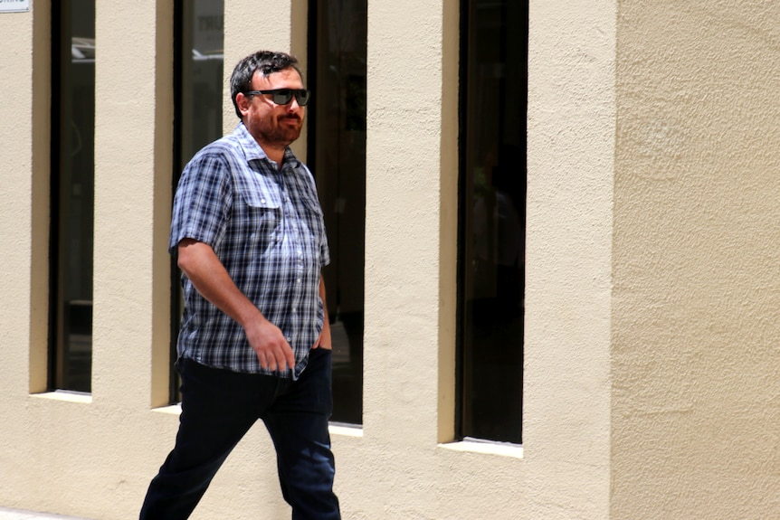 A man in a checked shirt and glasses walking outside a courthouse