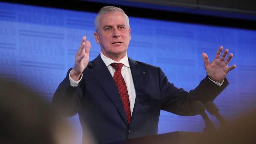 McCormack is giving a speech, standing behind a lectern, both arms are raised.