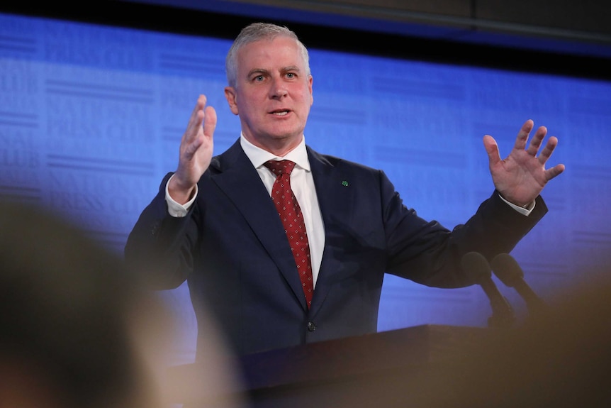 McCormack is giving a speech, standing behind a lectern, both arms are raised.