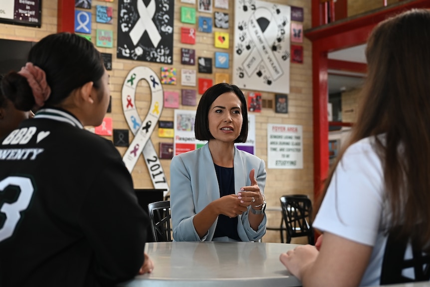 a woman sitting at a school desk talking to two high school students