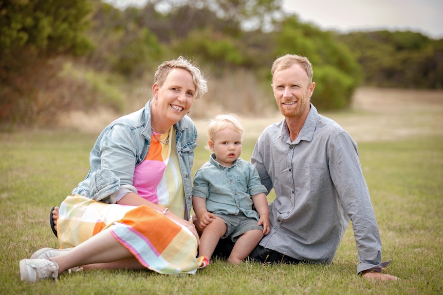 A family photo of a woman with her husband and baby sitting on the grass on a sunny day.