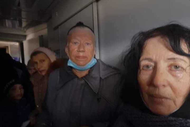 three women with sombre expressions look into the camera while standing on a train