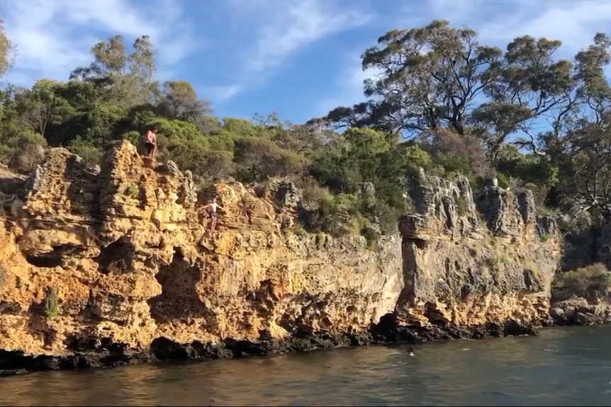 A man stands with his arms out above the water on rocks metres above the surface.