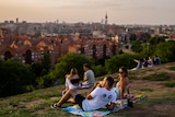 A group of people lounge on a picnic rug on the top of a green hill overlooking Madrid