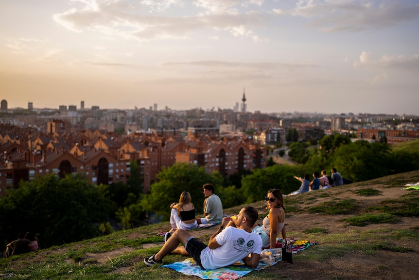 A group of people lounge on a picnic rug on the top of a green hill overlooking Madrid