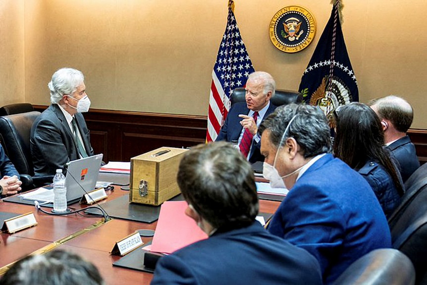 Joe Biden sits at the head of a conference table surrounded by colleagues wearing masks, with a US flag behind him