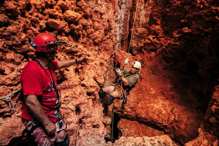 A man with red safety helmet and climbing gear supervises another man abseiling down red rocky cave terrain.