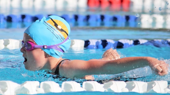 A girl is swimming butterfly with her arms extended back in a pool. She is wearing pink goggles and a blue swimming cap.