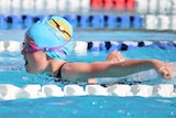 A girl is swimming butterfly with her arms extended back in a pool. She is wearing pink goggles and a blue swimming cap.