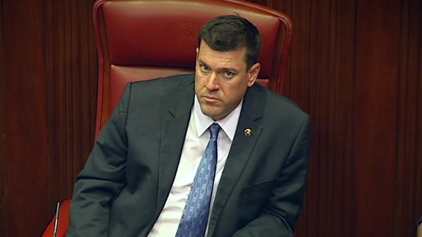 A man with a suit and tie sits in a chair in a parliament chamber