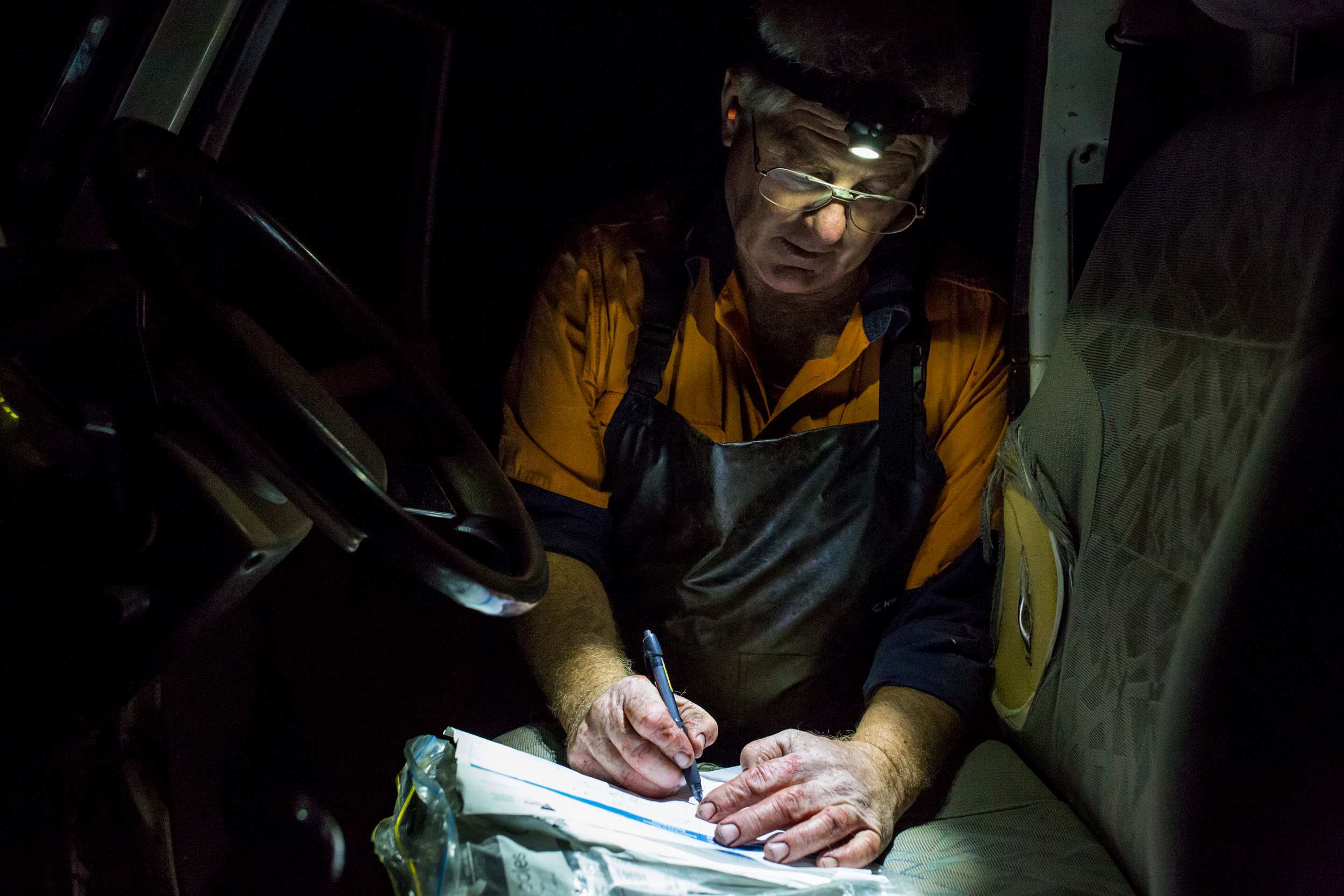 With an ear plug visible in one ear and red-stained hands, Glenn Cole does paperwork on the driver's seat of his truck.