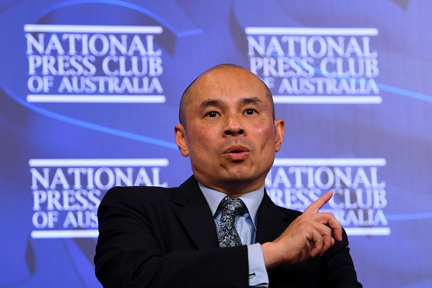 A man in a suit points over his left shoulder with his right index finger as he stands before a blue banner.