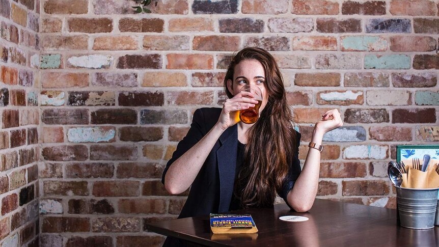 Colour photograph of woman drinking a beer in front of a brick wall, with her book on the table.