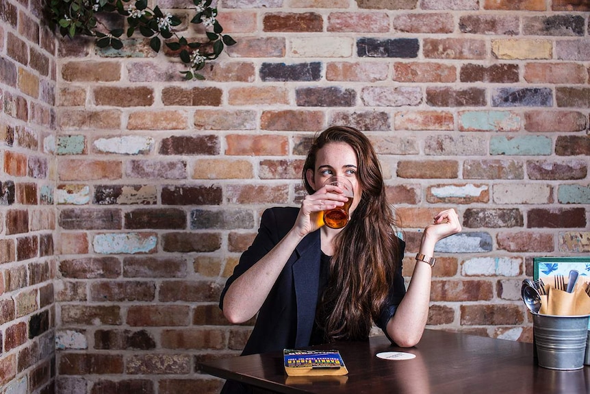 Colour photograph of woman drinking a beer in front of a brick wall, with her book on the table.