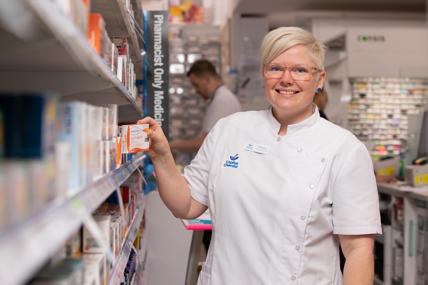A woman in a pharmacist uniform smiles as she fixes shelves.