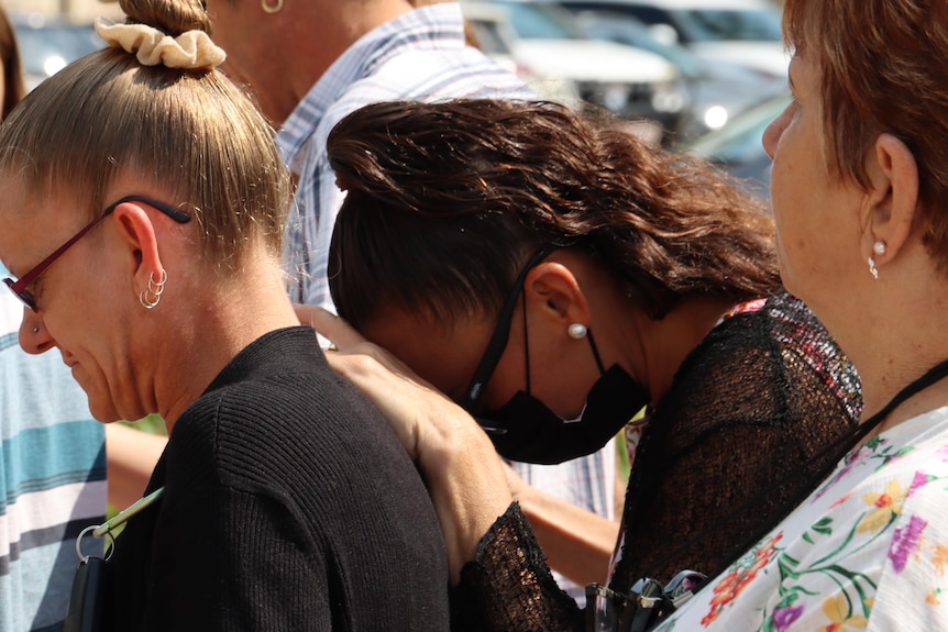 A woman visibly upset and wearing a black facemask rests her head on the shoulder of the person in front of her