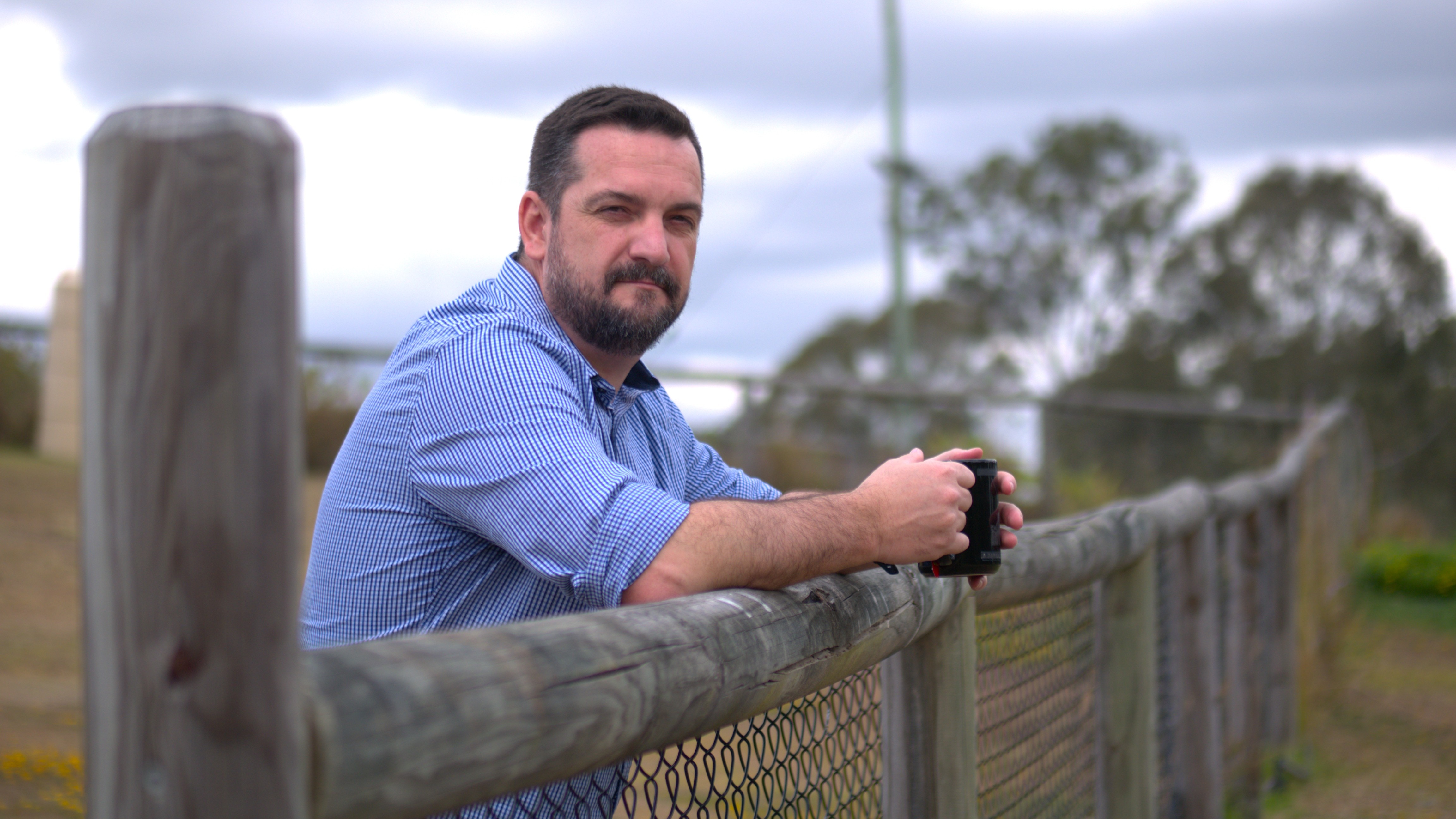 A man in a blue button up leans against a fence looking serious.