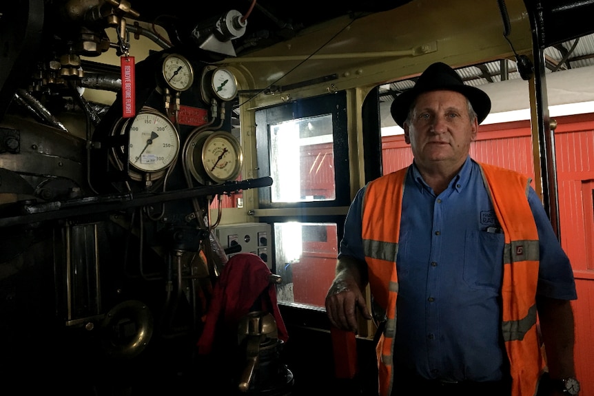A man wearing an orange high-vis vest stands beside gauges in a steam train cabin.