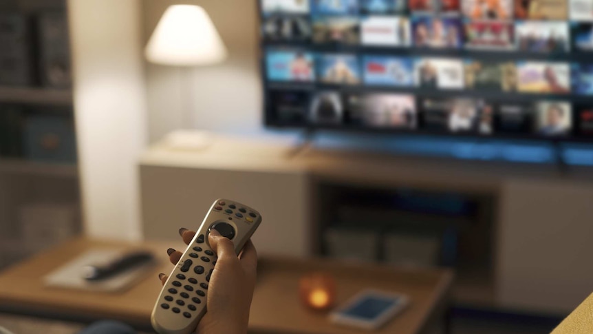 A hand with a remote control in front of a television screen showing a streaming service.