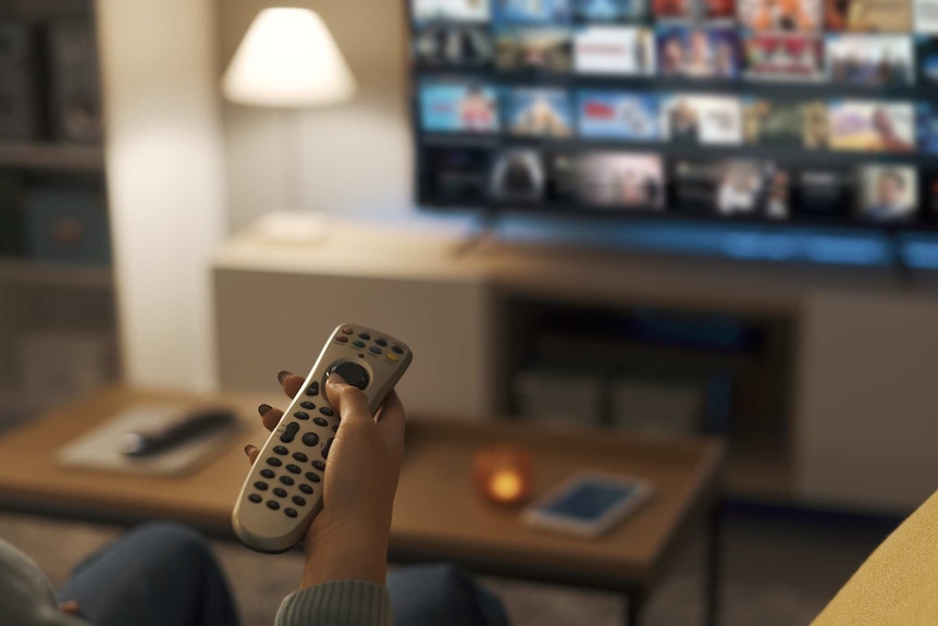 A hand with a remote control in front of a television screen showing a streaming service.