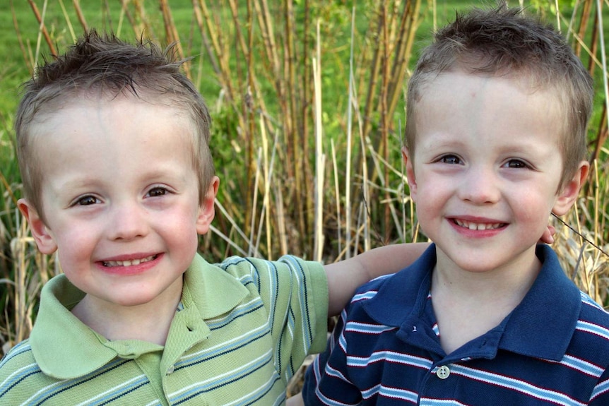Young identical twin boys smile into the camera. One is wearing a green t-shirt, and the other is wearing a blue t-shirt.