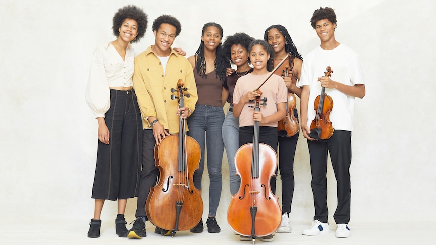 The seven Kanneh-Mason siblings stand in a row, some with their instruments, against a white background.