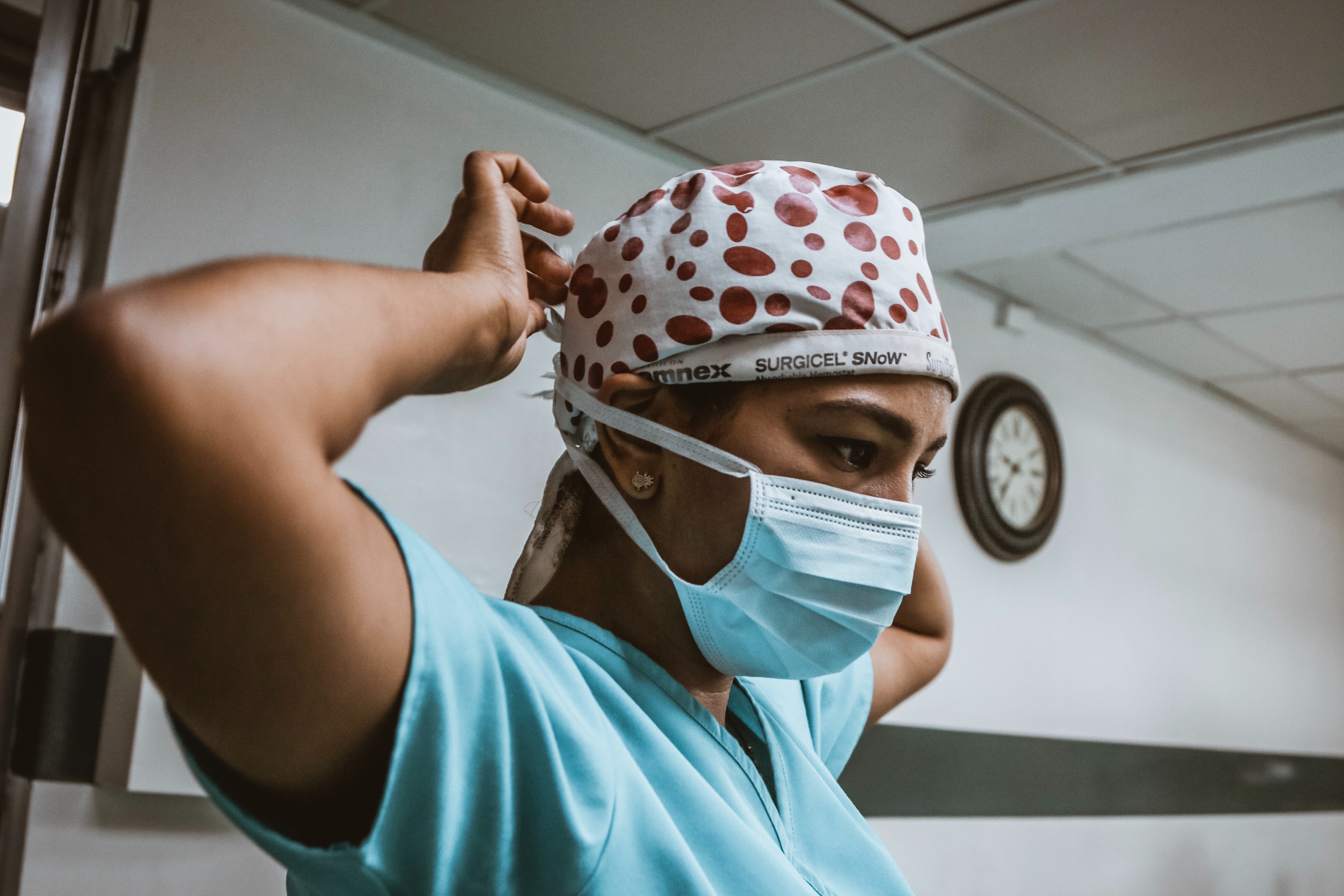 A woman with dark hair and light brown skin wearing blue scrubs and a spotted cap puts on a face mask.