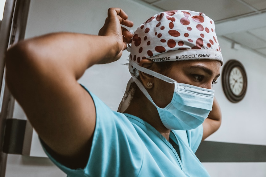 A woman with dark hair and light brown skin wearing blue scrubs and a spotted cap puts on a face mask.