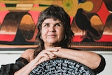 A 50-something Aboriginal woman with a mullet sits with her arms resting on a circular sculpture, handpainted with words