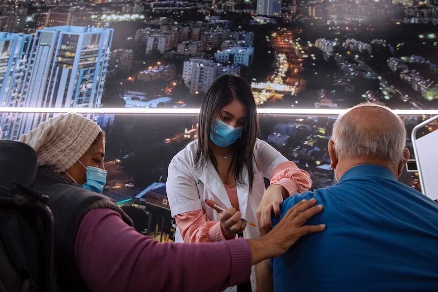 A woman in a face mask holds a needle to a man's arm