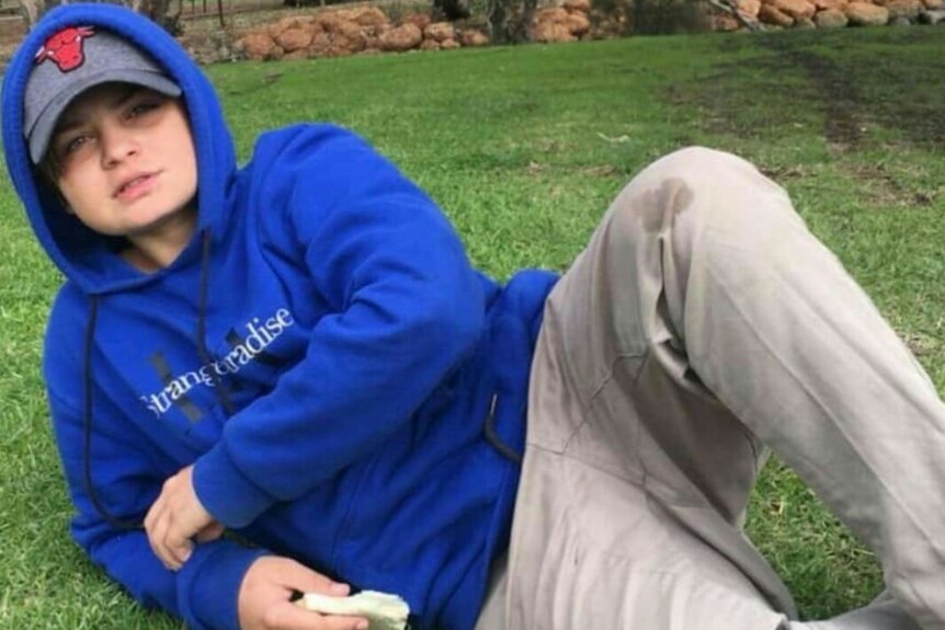 A young boy in a blue hoodie lies on the grass in a park.