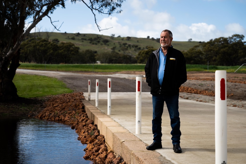 A man with a weathered face stands on a concrete pass looking over a creek on a sunny green acre lined with trees.