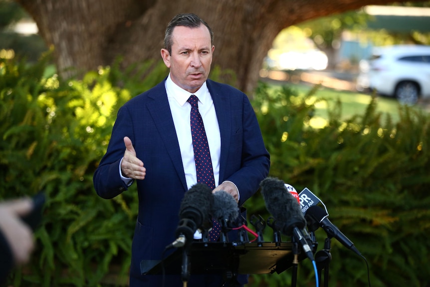 A mid-shot of WA Premier Mark McGowan speaking outdoors at a media conference with his right arm raised by his side.