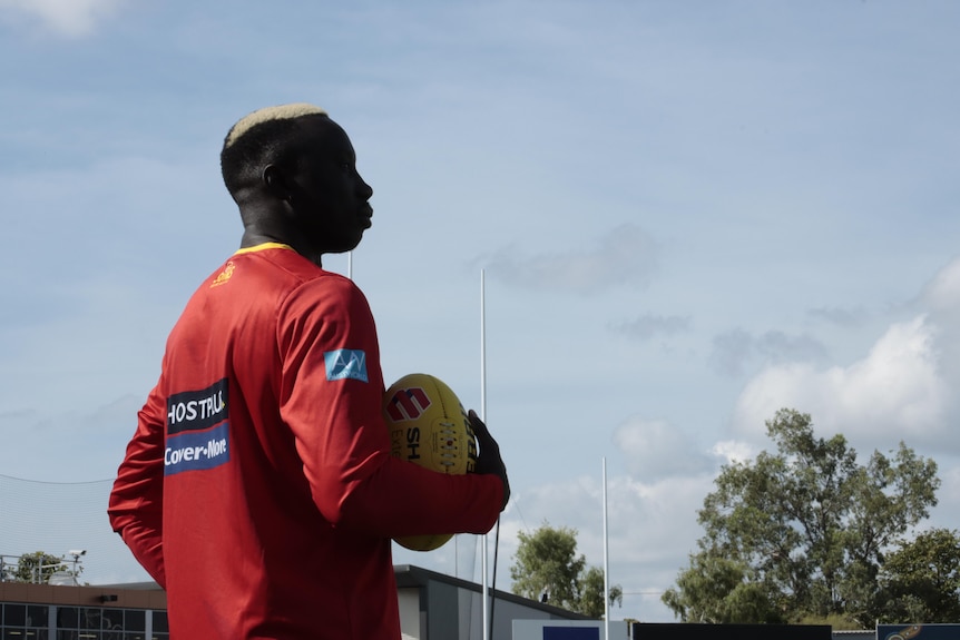 A man in a Gold Coast Suns shirt looks seriously off camera while holding a football.