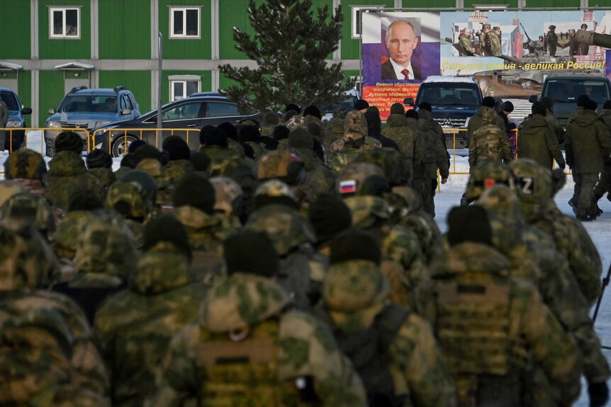 Dozens of Russian reservists in khaki military uniform, marching across snow toward a banner of Vladimir Putin.