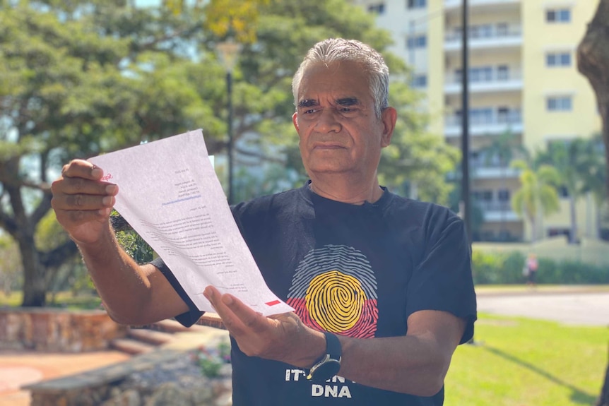 A man in a t shirt holds a letter in front of him and reads.