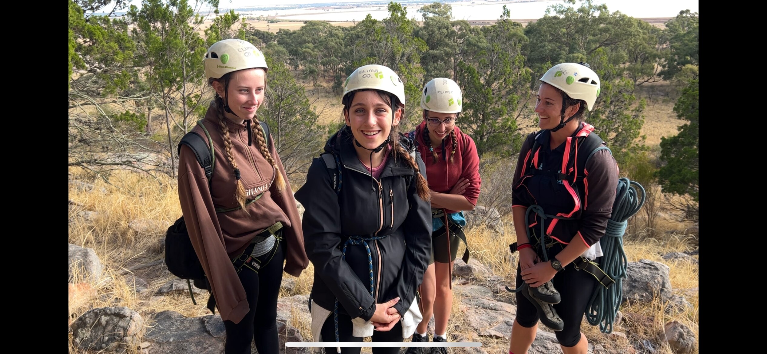 three girls and their teacher all wearing white helmets, carry climbing ropes smiling at the bast of a mountain 