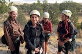 Three girls and their teacher all wearing white helmets, carry climbing ropes smiling at the base of a mountain.