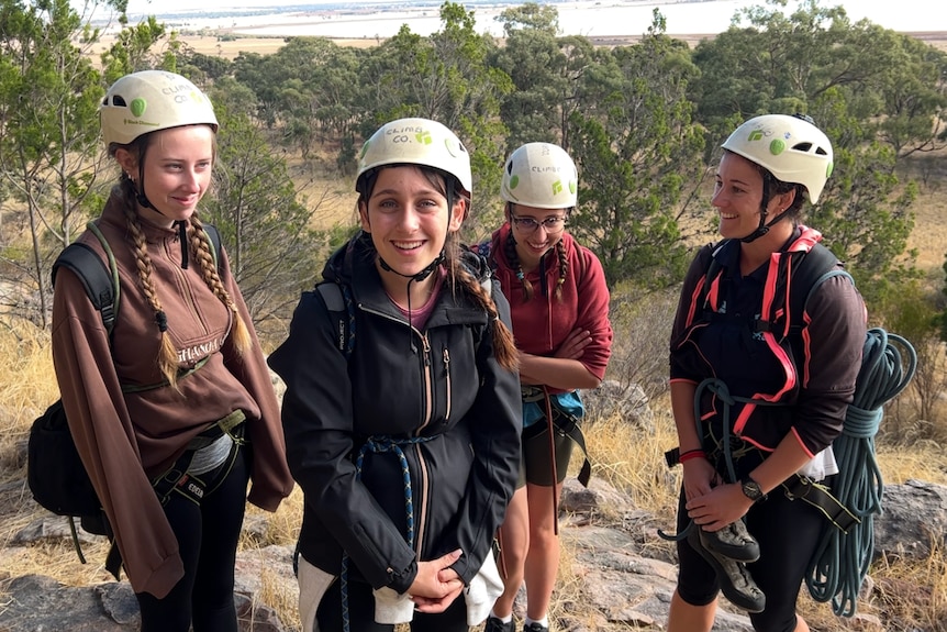 three girls and their teacher all wearing white helmets, carry climbing ropes smiling at the bast of a mountain 