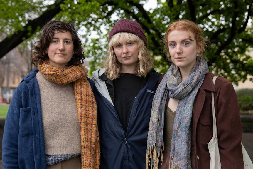 Three women stand together, looking seriously at the camera.