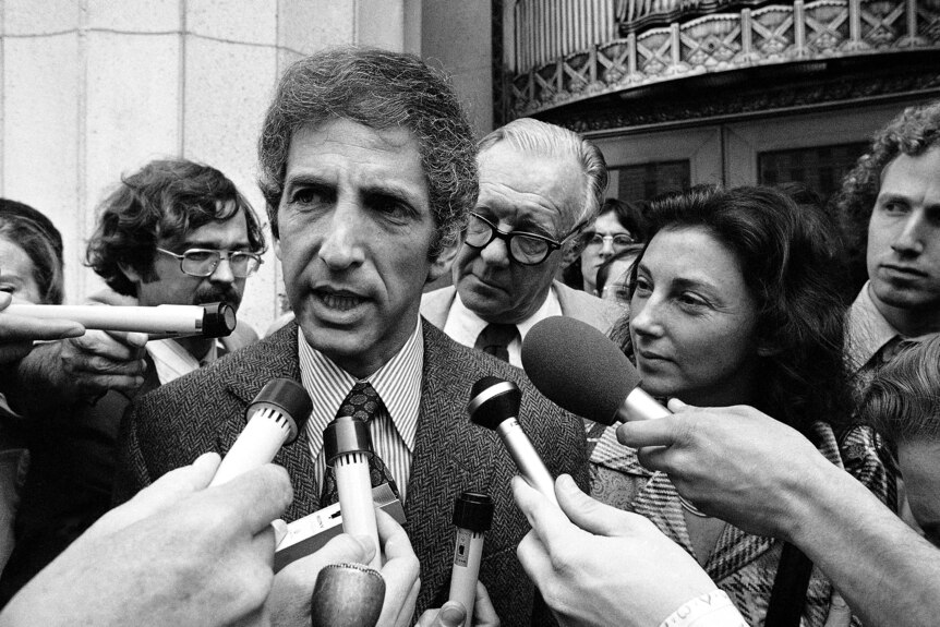 Black and white photo of Daniel Ellsberg speaking into press microphones in front of a building. He is surrounded by reporters.